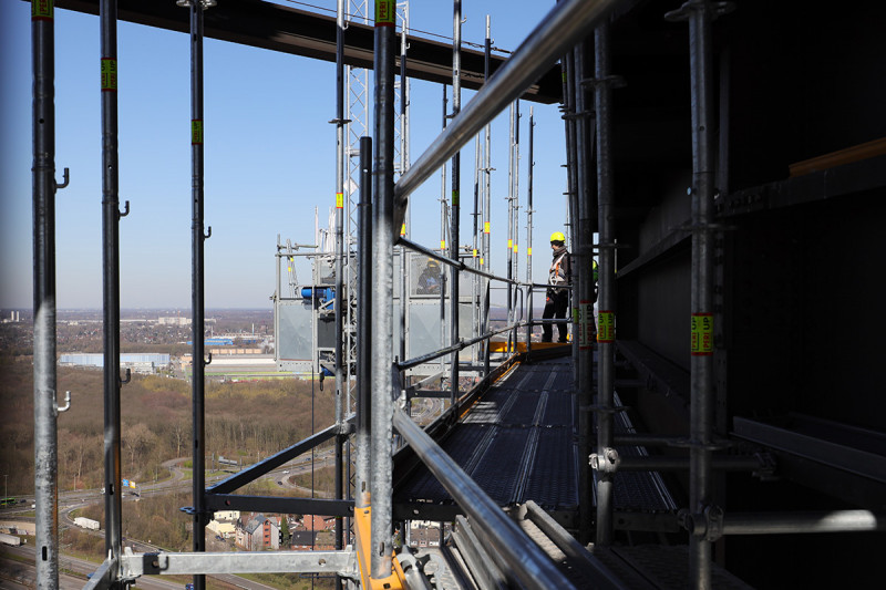 Ein Gerüstbauer auf dem Fassadengerüst des Gasometers unterhält sich mit einem Kollegen, der einen mobilen Aufzug bedient.