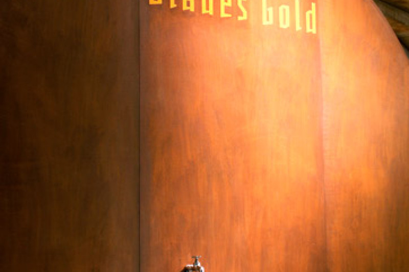 Großes halbrundes Fass mit goldener Blaues Gold Aufschrift. Darunter ein geöffneter Wasserhahn mit einem Einer der das Wasser auffängt.