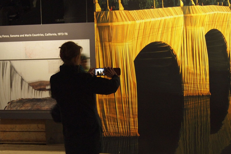 Verschwommenes Foto von einer im Vordergrund stehenden Person, die mit dem Handy ein großes Bild von einer gelben Brücke – Das goldene Leuchten der verhüllten Pont Neuf – fotografiert.